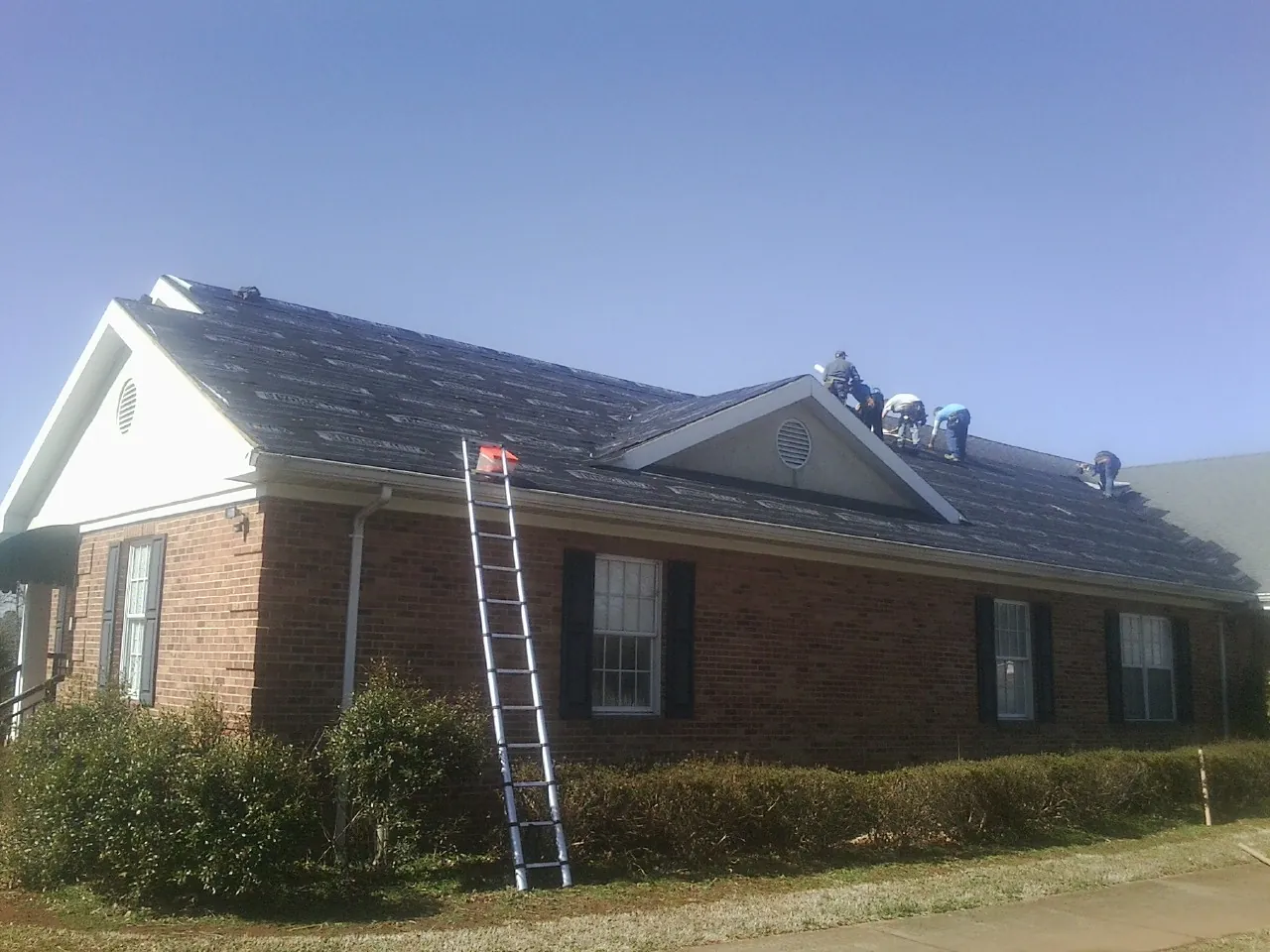 Crew tearing shingles off a brick ranch roof with a ladder set — Metal Roof Repair work by our crew in Orange City