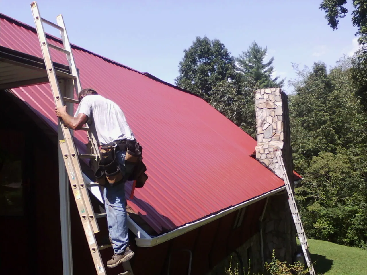 Roofer on a ladder beside a red metal roof and stone chimney — Soffit & Fascia Repair by Roofing Brothers in Orange City